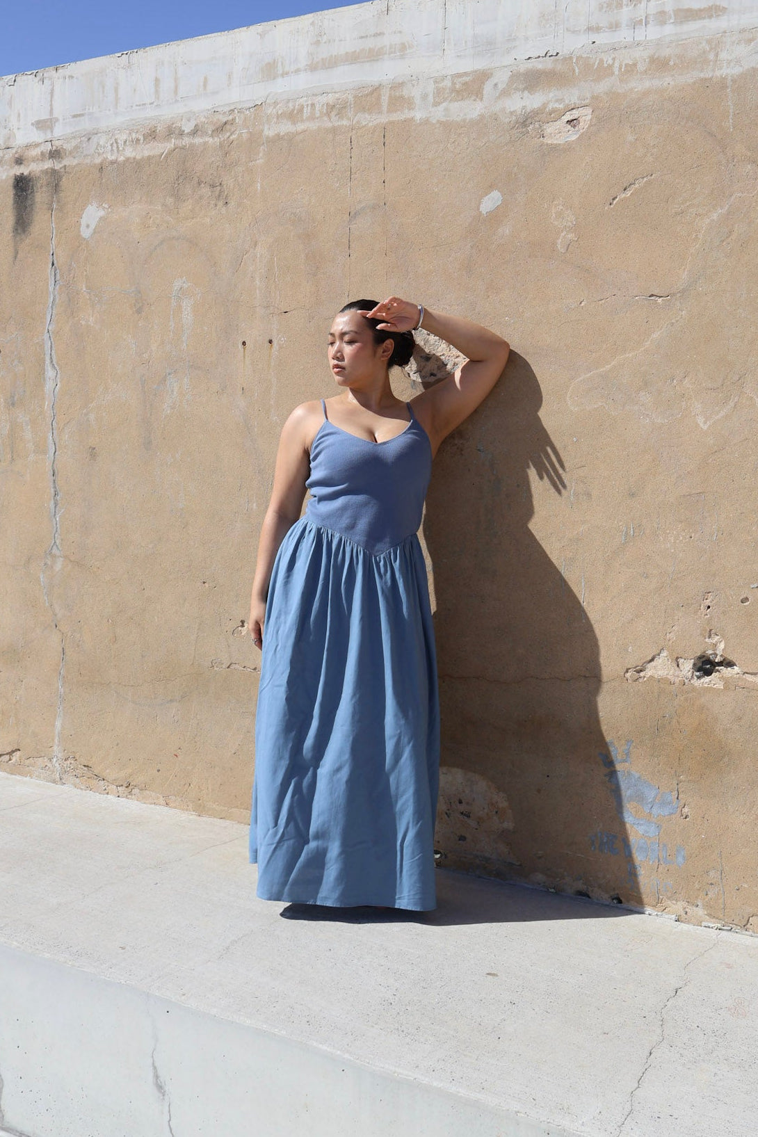 Woman in a blue dress standing against a beige wall with a clear blue sky.