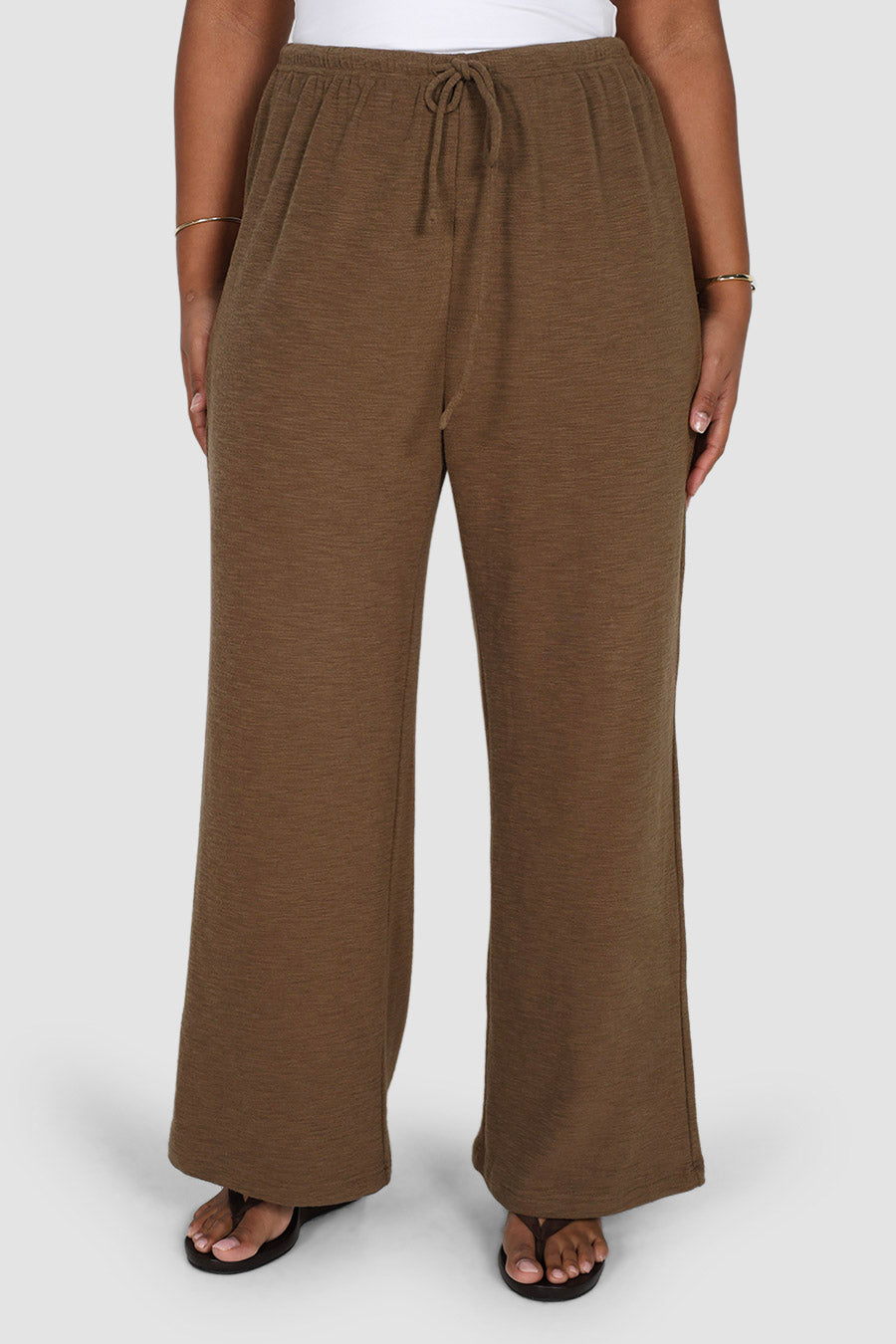 A person wearing loose, wide-leg drawstring pants in a medium brown color, paired with black flip-flops and a white top, shown against a light gray studio background.