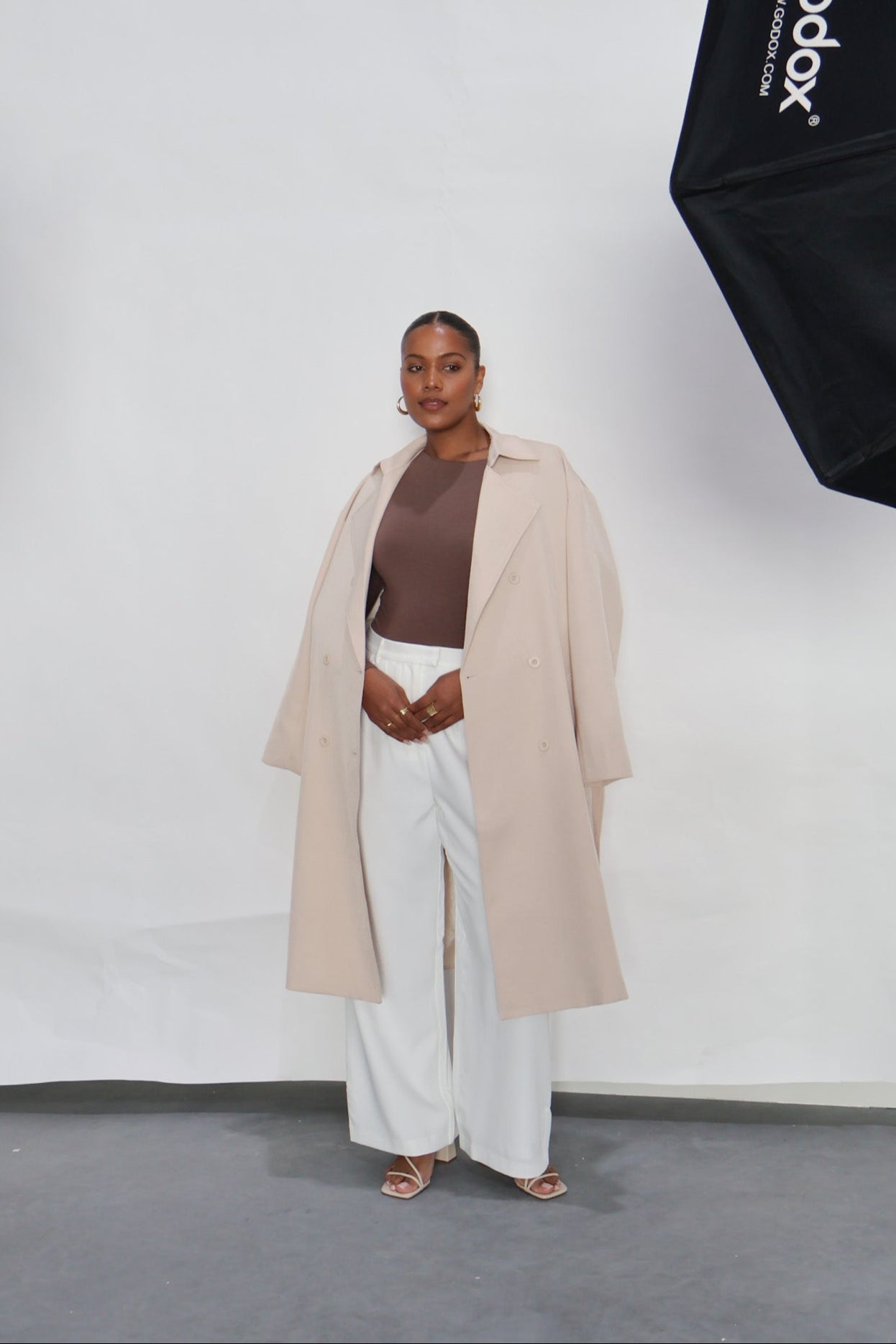 Person wearing a beige trench coat, brown top, and white pants in front of a white background with a Godox light stand.