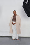 Person wearing a beige trench coat, brown top, and white pants in front of a white background with a Godox light stand.